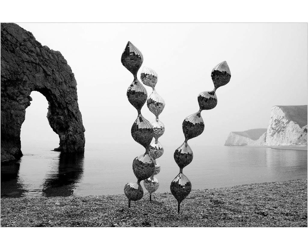An archived, silver mosaiced sculpture by Rebecca Newnham called Seedlings in black and white on a pebble beach. In the background is Durdle Door and a cloudy seascape. Photographed by David Bird.