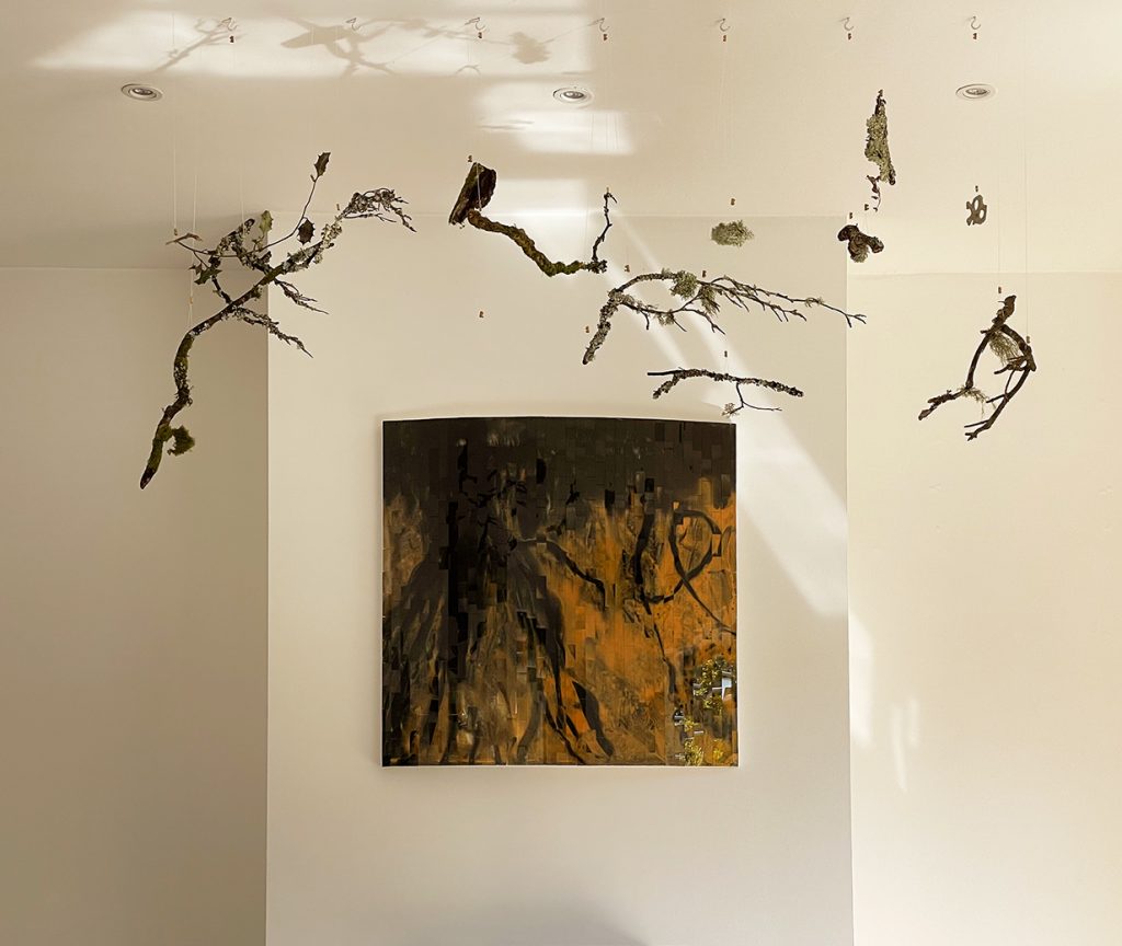 An archive sculptural wall panel by Rebecca Newnham to represent trees, lichen, and moss. It is black and gold, with dark streaks in the mosaic enamel. Above the piece are suspended branches with lichen growing on them photographed by David Bird.