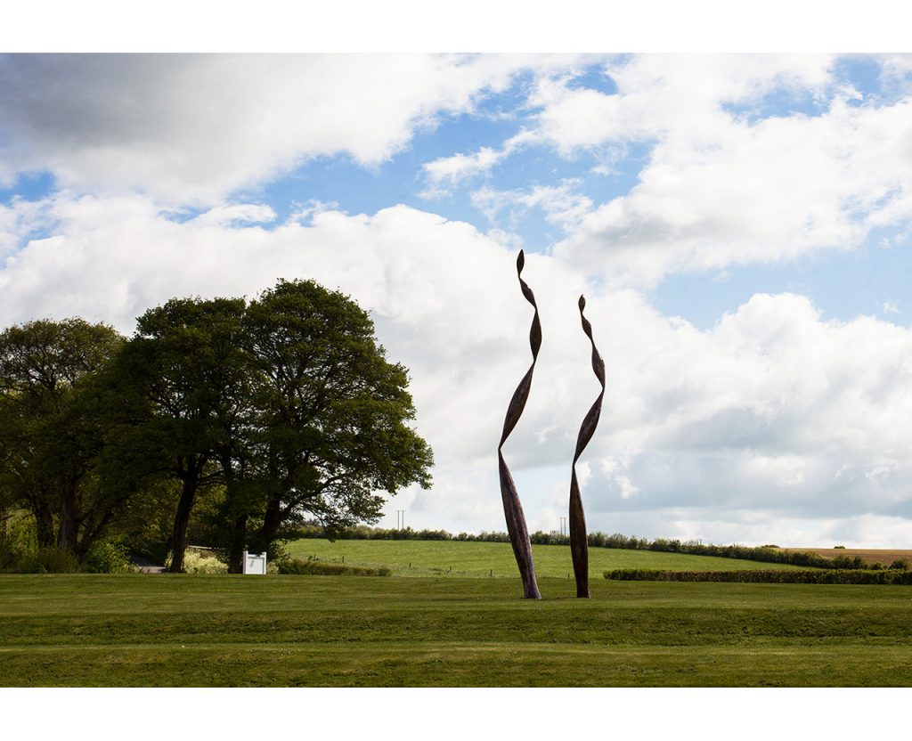 Two tall, archived red mosaic sculptures by Rebecca Newnham called Quercus standing in a green field. Photographed by David Bird.