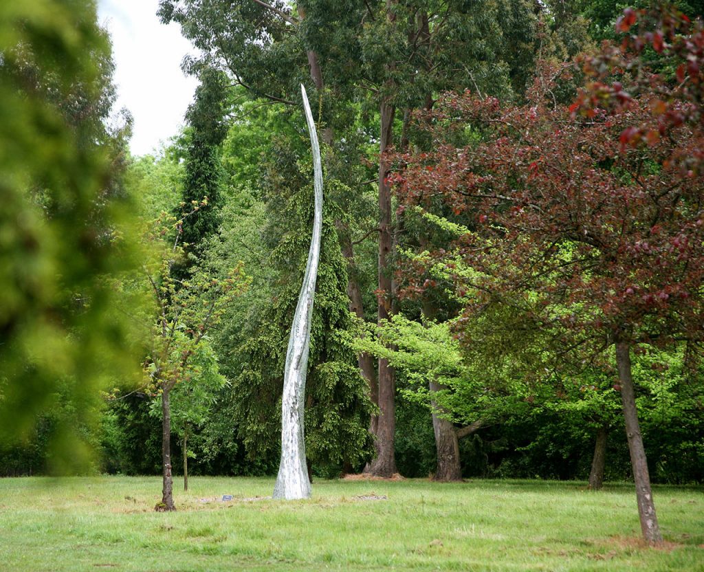 A tall, archived sculpture called Reed II by Rebecca Newnham mosaiced in silver enameled glass and standing among trees in early summer. Photographed by David Bird.